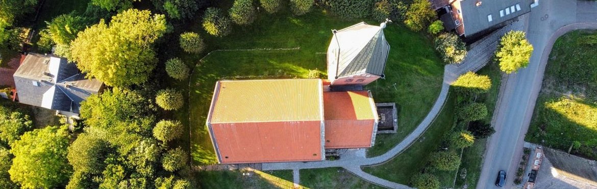 Eine Luftaufnahme zeigt das Dach einer Kirche, rundherum sind grüne Baume und rechts Wege und STraße zu sehen