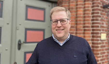 Ein Mann mit dunklem Pullover, Brille und blonden Haaren lächelt in die Kamera