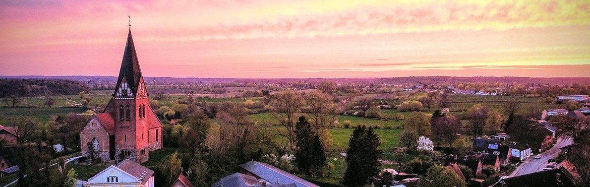 links sieht man eine Kirche in Luftaufnahme, ebenso die Dächer von einem Ort, der Himmel ist von lila-rot bis gelb