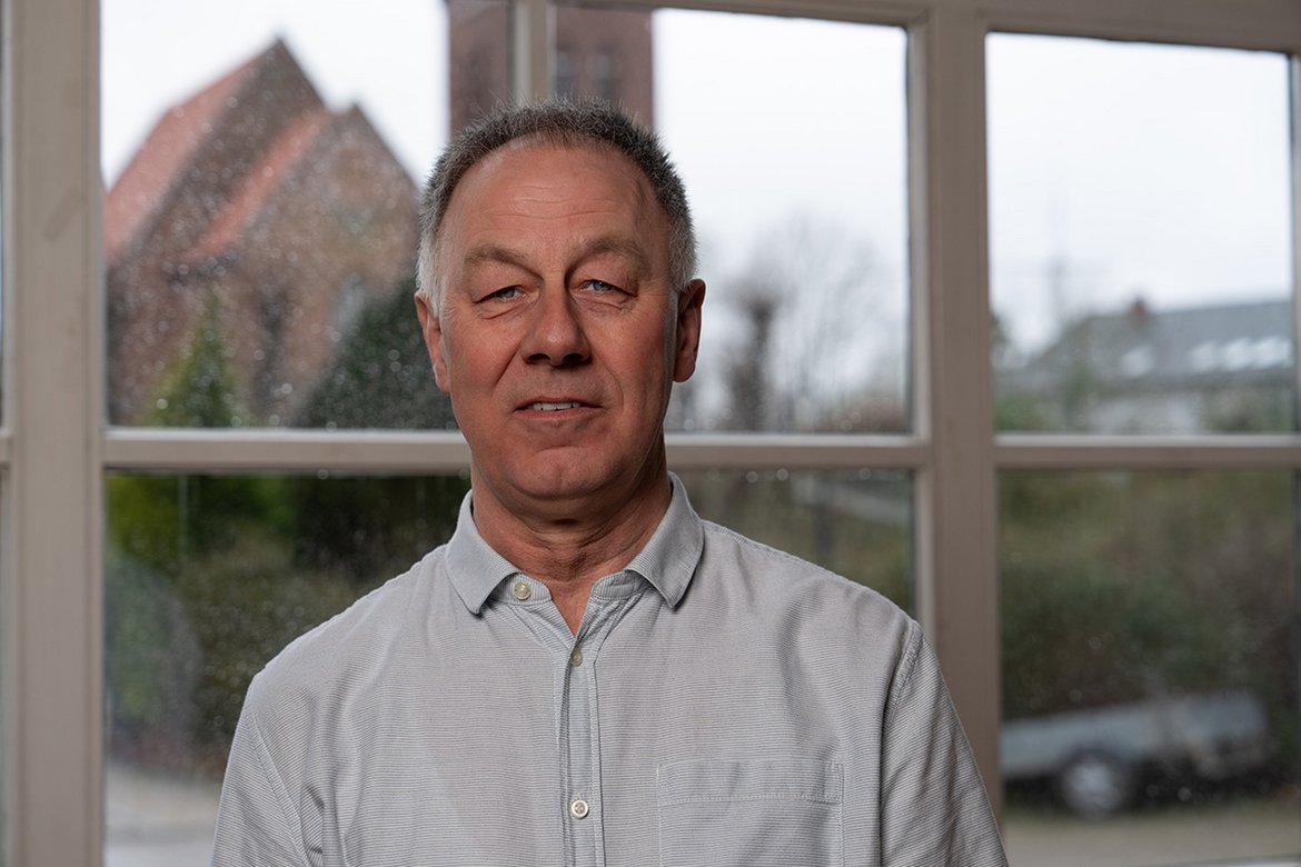 Rainer Rexin steht vor einem Fenster, im Hintergrund ist die Kirche Sandesneben zu sehen | Er trägt ein helles Hemd und lächelt in die Kamera