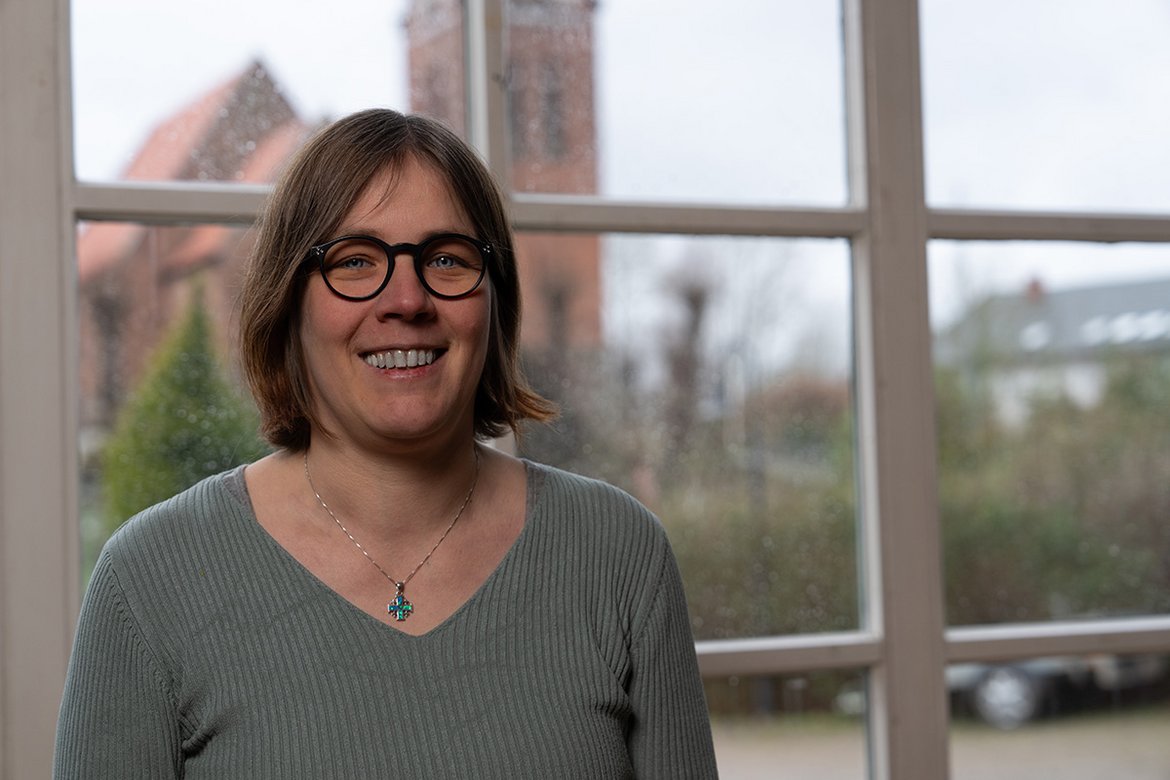 Doris Pfeifer lächelt in die Kamera, während sie vor einem Fenster steht. Im Hintergrund ist die Kirche Sandesneben zu sehen