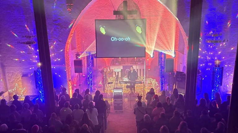 In einer vollen Kirche ist es dunkel. Der Altarraum ist umgebaut, es stehen dort ein Mann am Keybiard und eine Frau im goldenen Kleid steht vor einem Mikro. Viele Scheinwerfer sind an. Mehrere Diskokugeln leuchten und blaues und rotes Licht lässt alles festlich aussehen 