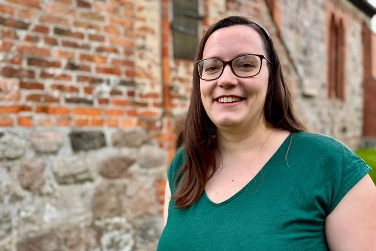 Junge Frau lächelt vor einer Backsteinkirche stehend in die Kamera.