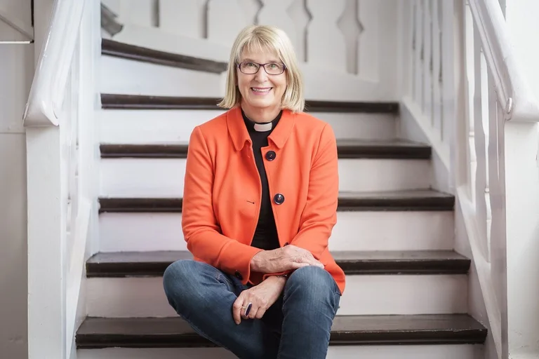 Eine Frau in Jeans, orangem Blazer und Collarhemd sitzt auf den Stufen einer weißen Holztreppe. Sie blickt freundlich in die Kamera.